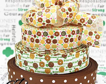 May include: A stack of brown, white, and green ribbon with a Girl Scout Cookie theme. The ribbon is decorated with images of Girl Scout Cookies and the words "Girl Scout Cookies".