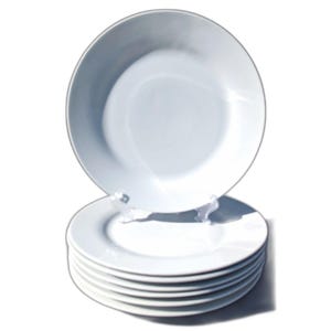 May include: A stack of six white ceramic plates, with a seventh plate displayed upright on a clear plastic stand. The plates have a simple, classic design, suitable for everyday dining or special occasions.