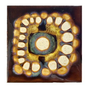 May include: Square ceramic tile with a unique abstract design. The tile features a dark brown background with a central circular shape surrounded by a ring of white and yellow circular shapes. The center circle is teal and gold.