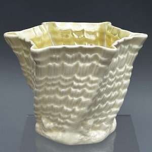 May include: A cream-colored ceramic vessel with a glossy, iridescent finish. The object features a distinct, wavy, ruffled rim and a textured, repeating shell-like pattern across its exterior surface, creating a dynamic, organic, and sculptural form.