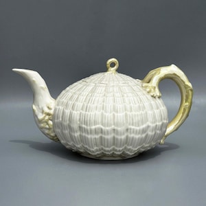 Op de afbeelding: Een witte keramische theepot met een getextureerd, geribbeld lichaam en een gebogen tuit. De handgreep en accenten zijn goudkleurig. Het deksel heeft een kleine lus om op te tillen. Een decoratief keukenartikel.