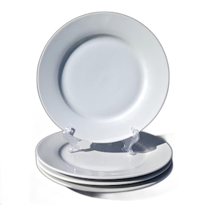 May include: A stack of four white ceramic plates, with a simple, clean design. One plate is displayed on a clear plastic stand. The plates have a wide rim and a slightly recessed center. The plates are suitable for everyday use.