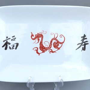 Könnte beinhalten: Eine rechteckige weiße Keramik-Servierplatte mit einem roten Drachen-Design und schwarzen chinesischen Schriftzeichen, die "Shou" und "Fu" lauten.