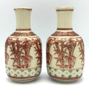May include: A pair of ceramic vases with a cream base and a red and gold bamboo design. The vases have a narrow neck with gold trim and a decorative band. The bottom features a white floral pattern with green accents. Each vase is approximately 15 cm tall.