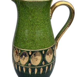 Op de afbeelding: Groene keramische kan met een getextureerd oppervlak en gouden accenten. De handgreep en rand zijn goudkleurig en een band met gouden medaillons versiert het onderste gedeelte. Een decoratief stuk voor het serveren van dranken.