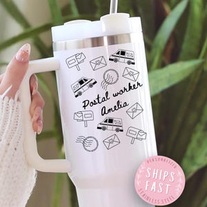 May include: White stainless steel tumbler with a handle, featuring black illustrations of mailboxes, mail trucks, and envelopes. The text "Postal worker Amelia" is printed on the tumbler. A pink sticker with the text "Ships Fast" is also visible.