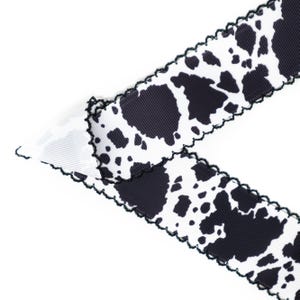 Peut inclure: Ruban blanc avec un motif de taches noires rappelant une vache. Le ruban a un bord festonn&eacute; avec une bordure noire. Le ruban est inclin&eacute; sur un fond blanc.