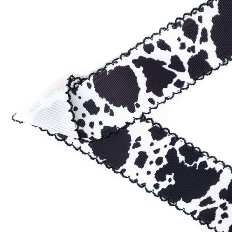 Peut inclure: Ruban blanc avec un motif de taches noires rappelant une vache. Le ruban a un bord festonn&eacute; avec une bordure noire. Le ruban est inclin&eacute; sur un fond blanc.