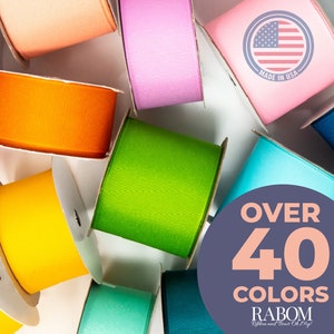 May include: A variety of colorful ribbon spools in different colors, including orange, yellow, green, blue, and pink. The spools are arranged in a circle around a sign that says "Over 40 Colors" and "Rabom Ribbon and Bow Co. Mfg."