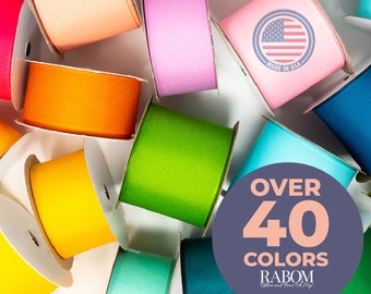 May include: A variety of colorful ribbon spools in different colors, including orange, yellow, green, blue, and pink. The spools are arranged in a circle around a sign that says "Over 40 Colors" and "Rabom Ribbon and Bow Co. Mfg."