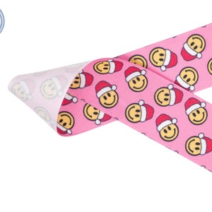 May include: Pink ribbon with a repeating pattern of yellow smiley faces wearing red Santa hats.
