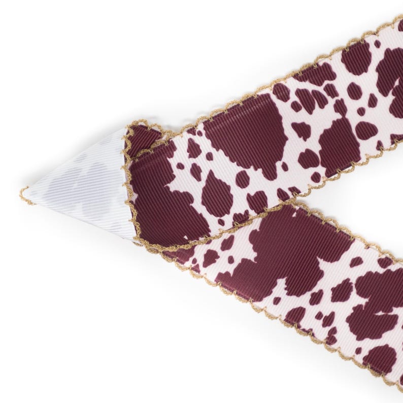 Peut inclure: Ruban d&eacute;coratif avec un motif de vache. Le ruban pr&eacute;sente un fond blanc avec des taches marron et un bord festonn&eacute; dor&eacute;. Le ruban est pli&eacute; sur lui-m&ecirc;me, r&eacute;v&eacute;lant le dessous blanc.