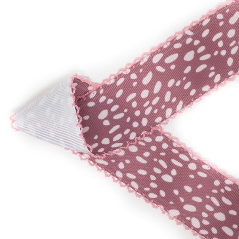 Peut inclure: Ruban d&eacute;coratif avec un fond mauve et des pois blancs. Le ruban a un bord festonn&eacute; rose et un dessous ray&eacute; blanc et gris. Le ruban est pli&eacute; sur lui-m&ecirc;me.