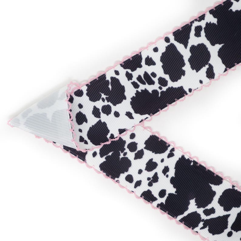 Peut inclure: Ruban blanc orn&eacute; d'un motif de taches noires rappelant une vache, bord&eacute; d'une finition festonn&eacute;e rose. Le ruban est pli&eacute;, r&eacute;v&eacute;lant un envers ray&eacute; blanc et gris. Id&eacute;al pour l'artisanat ou l'emballage cadeau, environ 2.5 cm de large.
