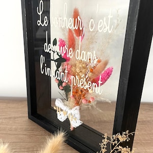 Peut inclure: Une boîte à ombre encadrée de noir avec un arrangement de fleurs séchées et une inscription blanche qui dit "Le bonheur, c'est de vivre dans l'instant présent".