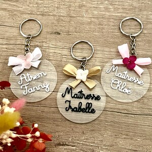 Peut inclure: Trois porte-clés en acrylique transparent avec des nœuds de différentes couleurs et un texte personnalisé. Les porte-clés comportent le texte "Atsem Fanny", "Maitresse Isabelle" et "Maitresse Chloé".