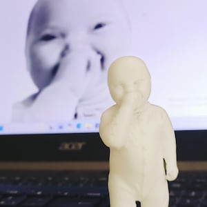 May include: A small, off-white figurine of a baby in a onesie, with one hand raised to its face. The background features a blurred image of a baby on a computer screen. The figurine is standing in front of a laptop keyboard.
