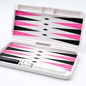 Könnte beinhalten: Offenes weißes Backgammon-Set mit einem rosa, schwarzen und weißen Dreiecksmuster. Das Set enthält schwarze und weiße Spielsteine, Würfel und einen schwarzen Würfelbecher. Das Spielbrett ist offen und spielbereit.