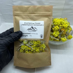 Op de afbeelding: Een bruine papieren zak met een doorzichtig venster, gevuld met gele Everlasting Flower (Helichrysum arenarium). Het etiket luidt "Herbs of Karpaly", geoogst in de zomer van 2025 en weegt 57g. Een kom met gedroogde gele bloemen staat op de achtergrond.