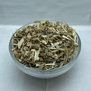 May include: A clear glass bowl filled with a heap of dried, shredded light brown and tan woody bark pieces. The texture of the botanical material is rough and fibrous, consisting of small, irregular slivers and fragments of plant matter.