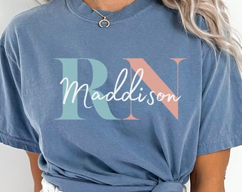 May include: A dusty blue t-shirt with the letters "RN" in teal and coral, and the name "Maddison" in a cursive font. The shirt is knotted at the waist. The model is wearing gray sweatpants.
