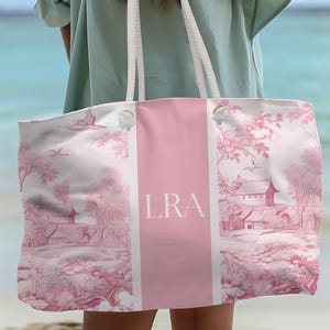 May include: A large pink and white tote bag with a pink stripe down the center. The bag has a white rope handle and features a pink toile pattern with a small house and trees. The bag is personalized with the initials "LRA" in white.