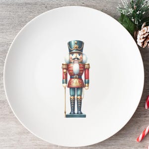 May include: A white ceramic plate featuring a colorful illustration of a traditional nutcracker soldier. The nutcracker is dressed in a red and blue uniform with gold accents. Christmas-themed decor includes a pine cone, berries, and candy canes.
