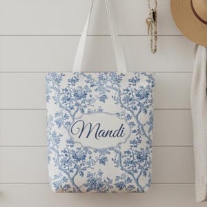 May include: A white tote bag with a blue floral pattern and white straps hangs on a wall. The bag features the name "Mandi" in a decorative font within a framed design. A straw hat and keys hang nearby.