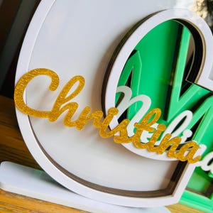 May include: A white, layered, heart-shaped frame with a gold glitter "Christina" nameplate.