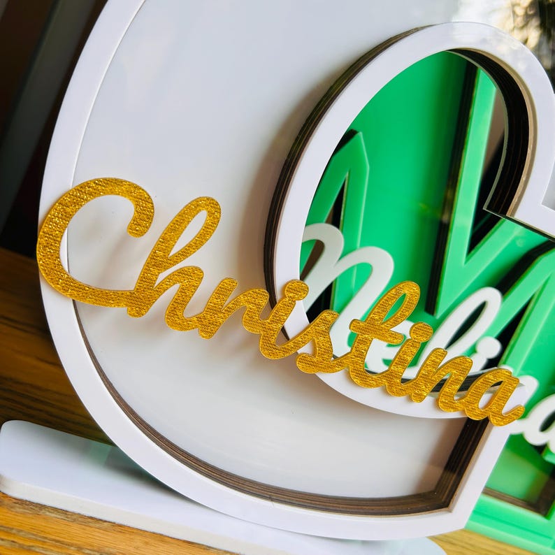 May include: A white, layered, heart-shaped frame with a gold glitter "Christina" nameplate.