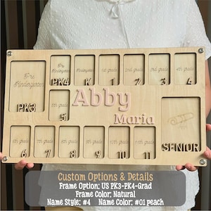 May include: A wooden school years photo display board with spaces for pre-kindergarten through 11th grade, plus a senior year slot. The name "Abby Maria" is in pink letters. The board is light-colored wood.