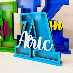 May include: A blue acrylic letter A with a black frame and a white name "Abric" inside. The letter is standing on a white surface.