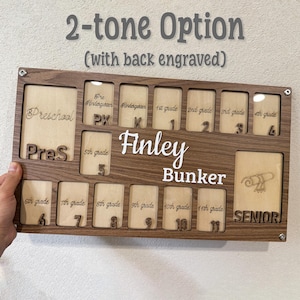 May include: A two-tone wooden school years display board with spaces for photos from preschool through senior year. The name "Finley Bunker" is displayed in white lettering. The text "2-tone Option" is at the top.