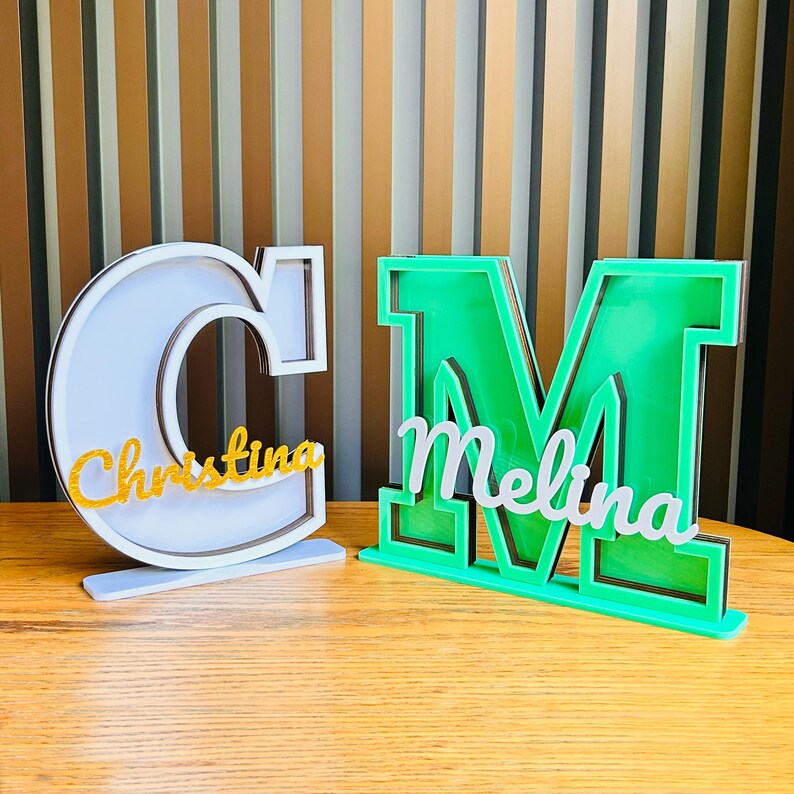 May include: Two wooden letters, a white "C" and a green "M", stand on a wooden surface. The letters are decorated with gold glitter and the names "Christina" and "Melina" are written on them.