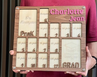 May include: Wooden school years photo display board with the name "Charlotte Jean" in pink. The board has slots for photos and grade levels from Pre-K to 12th grade, with a graduation cap and diploma design.