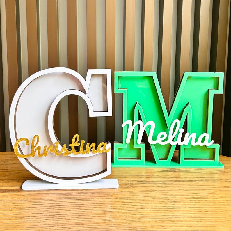 May include: Two wooden letters, a white "C" with the name "Christina" in gold glitter letters inside and a green "M" with the name "Melina" in white letters inside.
