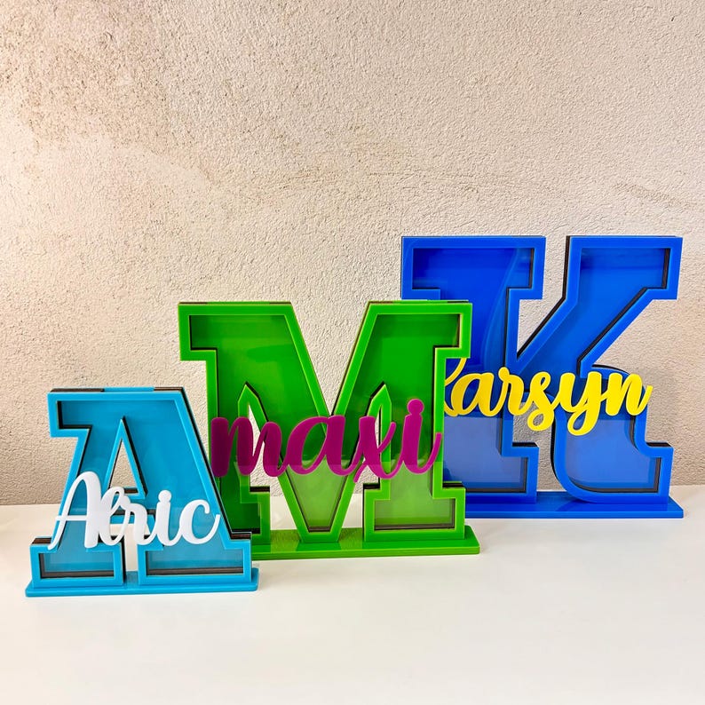 May include: Three colorful, freestanding letters, A, M, and K, are made of plastic and have a slot in the front. The letters are painted in different colors: blue, green, and blue. The letters have names inside: Abic, maxi, and karsyn.