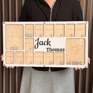 Peut inclure: Un tableau de graduation personnalisé en bois avec le nom "Jack Thomas" en écriture cursive. Le tableau comporte des emplacements pour chaque niveau scolaire, de la maternelle à la terminale, et est encadré en blanc.