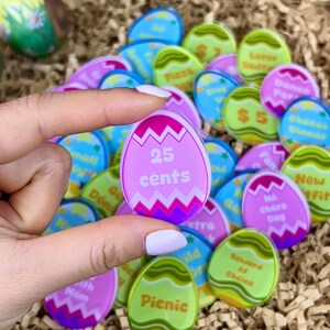 May include: A collection of colorful, egg-shaped tokens with various designs and text. One token, held in a hand, displays "25 cents" in pink. Other tokens feature phrases like "Pizza," "Picnic," and "No Chore Day."