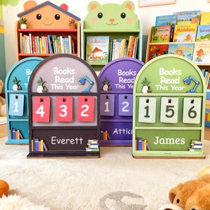 May include: Wooden book counters in blue, gray, purple, and green, each with a different number displayed. The counters read "Books Read This Year" and have names like "Everett" and "James" at the bottom. Bookshelves are in the background.