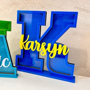 May include: Blue acrylic letter K with a yellow name "Karsyn" inside. The letter is freestanding and has a base.