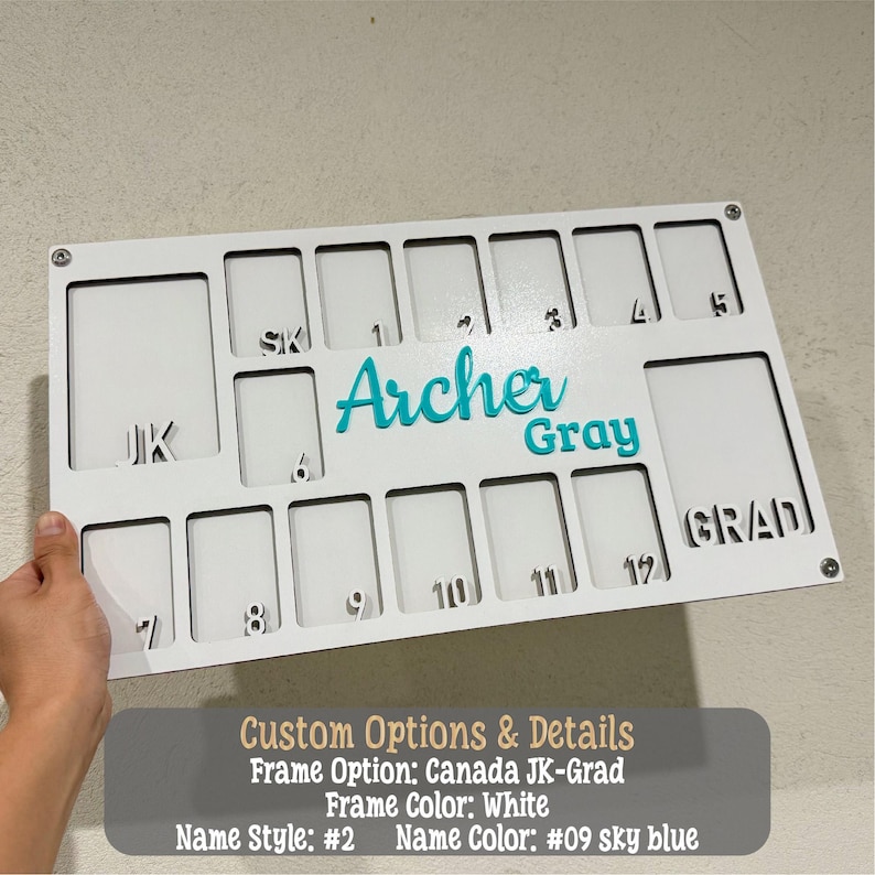 May include: A white wooden graduation board with cut-out photo slots and the name "Archer Gray" in teal. The board includes the text "GRAD" and numbers 1-12. The board is held by a hand.