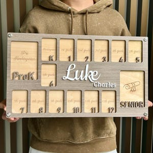 May include: A wooden photo frame with 12 picture slots for each grade of school, from Pre-K to Senior year. The frame is personalized with the name "Luke Charles" and the text "Pre-K", "Kindergarten", "1st grade", "2nd grade", "3rd grade", "4th grade", "5th grade", "6th grade", "7th grade", "8th grade", "9th grade", "10th grade", "11th grade", "12th grade", and "Senior".