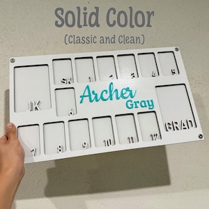 May include: A white wooden board with cut-out rectangular slots and the words "Solid Color" and "(Classic and Clean)" in gray. The name "Archer Gray" is in teal. Numbers and letters are cut out for personalization.