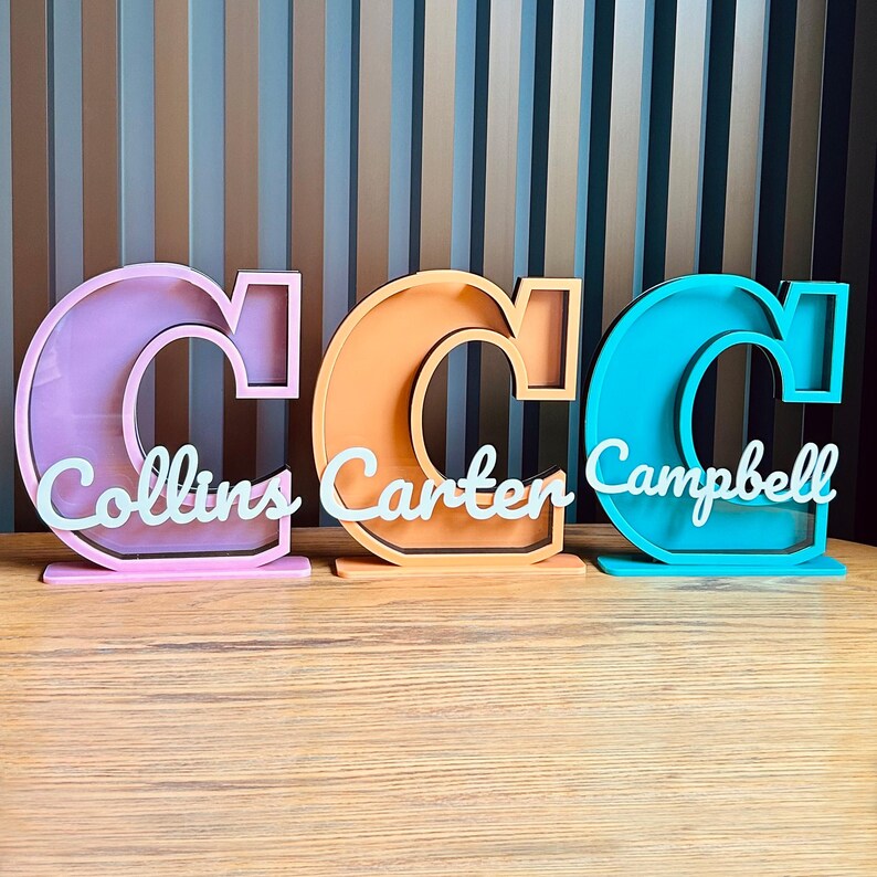 May include: Three wooden piggy banks shaped like the letter C, each painted in a different color: pink, orange, and blue. The names "Collins", "Carter", and "Campbell" are written on the front of each piggy bank.