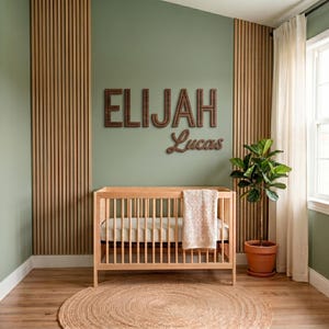 Op de afbeelding: Een houten wieg met een lichtgekleurd matras en een crèmekleurige deken, geplaatst onder aan de muur bevestigde houten letters die ELIJAH in blokletters en Lucas in cursief spellen, tegen een saliegroene muur met verticale houten latten.