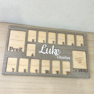 May include: A wooden school years photo display board with spaces for photos from Pre-K to Senior year. The board is gray with a wood grain pattern, and the name "Luke Charles" is in white script.