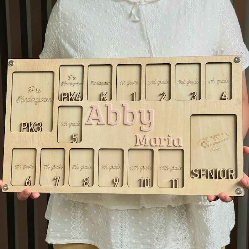 May include: A wooden school years photo display board with spaces for photos from Pre-Kindergarten to Senior year. The name "Abby Maria" is in pink letters. The board is held by a person.