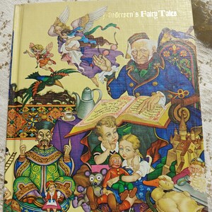 May include: A hardcover book titled "Andersen's Fairy Tales" with a colorful illustration. The cover features a detailed painting of various fairy tale characters, including a man reading a book, children, and fantastical creatures.