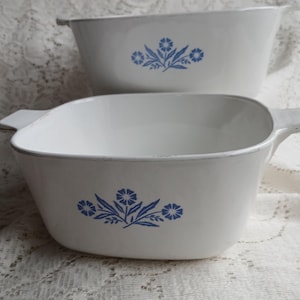 May include: Two white casserole dishes with blue floral designs. Each dish has a rectangular shape with rounded corners and a handle on each side. The dishes are stacked on top of each other.